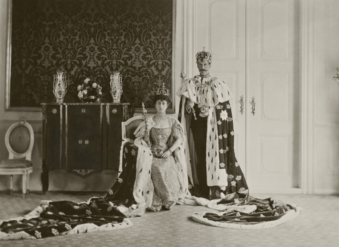 Kong Haakon og Dronning Maud med riksregalier og kroningskapper, fotografert i Stiftsgården 22. juni 1906. Kappen i utstillingen ble opprinnelig laget til Dronning Joséphine, og er 390 cm lang. Den er fôret med silke og har to dekorerte bånd sydd på fronten. Foto: De kongelige samlinger